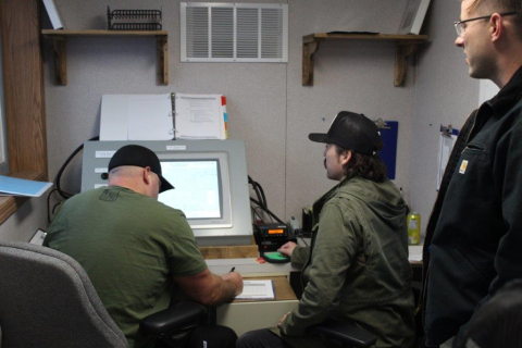 Three employees standing and sitting by a computer screen and monitoring