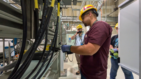 Three employees in yellow hard hats, one employee holds a tool and works with it