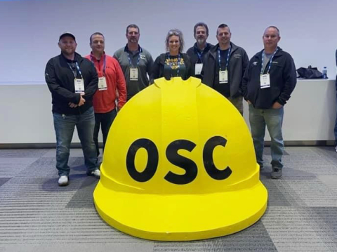 A group of workers stand around a large yellow hard hat with the letters "OSC" on it