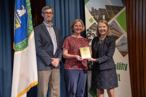 Three people stand on a stage and smile for a picture, two of the people hold an award plaque 