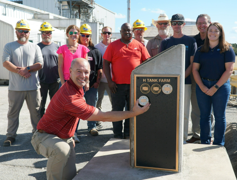 A large group of people pose for a group picture in front of an outside plaque