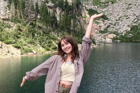 A woman standing in front of a lake surrounded by mountains. She is smiling at the camera.