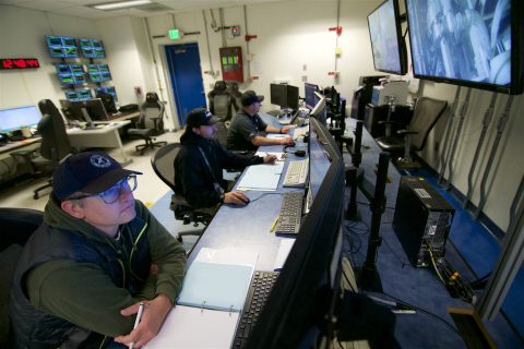 Three employees sit at a long desk with lots of computers and screens on the desk and wall in front of them