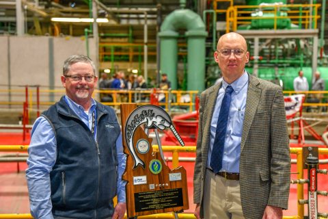Two men pose for a photo beside an award plaque inside a laboratory building