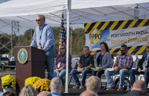 A man stands at a podium on a stage with people sitting behind him. He speaks to a crowd