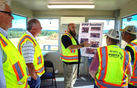 A group of people in protective gear stand in a room and listen to a man speak and present a poster