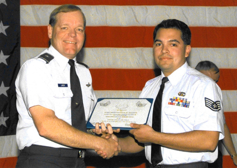 Two men in military uniforms shake hands and hold a certificate while standing in front of an American flag