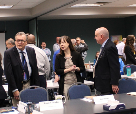 Three professionals stand in a room full of people and speak to each other
