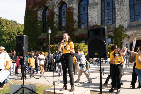 Emilie Lozier stands in front of an academic building with many people in the background and surrounding her and speaks into a microphone. 