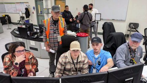 A group of five people sitting and standing around a monitor in a control room as they watch the screen