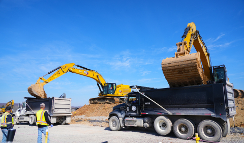 Two work machine vehicles scoop dirt into the back of two large trucks, workers stand around in yellow protective vests