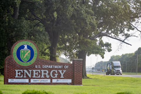 A brown sign with white words and the DOE seal beside a road with a truck driving on it