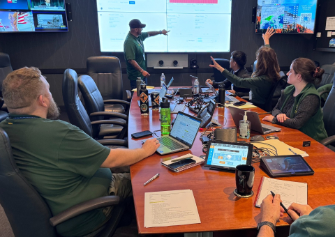 People sit around a conference table and look at the screen at the front of a room where a man stands and points at the screen