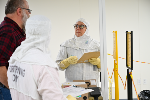 An employee in a white safety suit holds a clipboard and looks at two other employees
