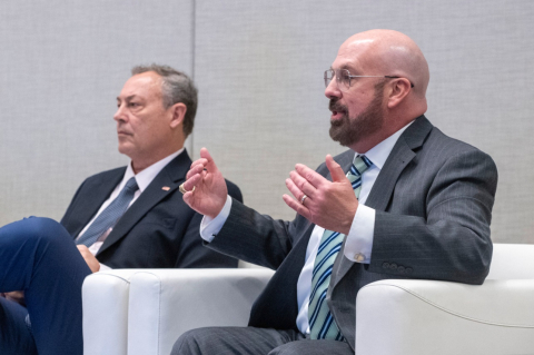 Two men sitting in white chairs, one is speaking and slightly raising both his arms