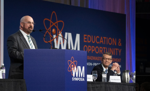 A man stands at a podium and delivers a speech, two other individuals sit at a table on the stage behind him