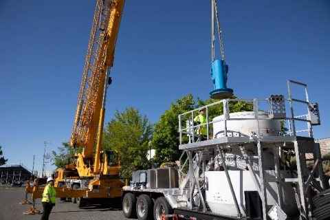 A crane lowers a blue cask into a container sitting on a flatbed trailer.