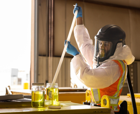 An employee in protective gear stirring a sample with a long object