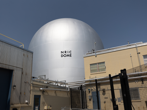 The NRIC DOME facility at INL, a tall, silver dome-shaped building against a blue sky.