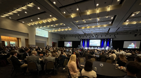 A large conference room with tables filled with people listening and watching the presentation on stage