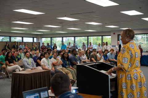 A woman at a podium speaking to a group of interns sitting in chairs in the crowd