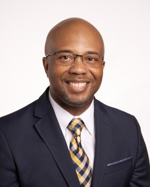 A portrait of a man in glasses and a black suit with a yellow and black tie