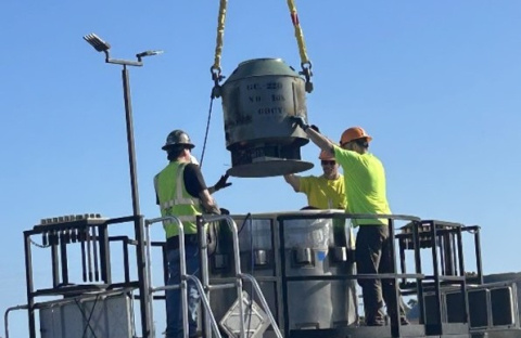 Two men in high-visibility clothing carefully lower a cesium irradiator into a transportation cask with the help of a crane.