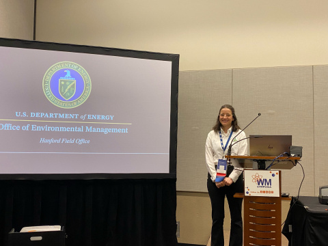 A professional posing for a picture by a podium and a large screen with the DOE EM seal on it