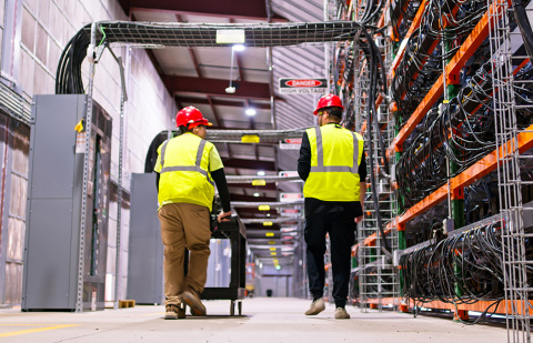 Two technicians wearing PPE walking past racks of equipment in a data center