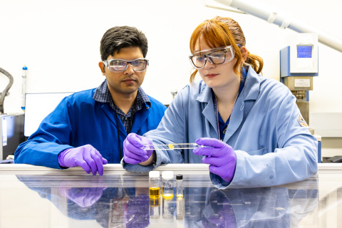 Mary Danielson, who is wearing a lab coat, goggles, and gloves, is holding two pieces of glass attached together with the yellow adhesive. She is leaning on a reflective lab bench with samples of the adhesive in small glass bottles in front of her. Anisur Rahman (who is also in lab equipment) is standing next to her, watching. 