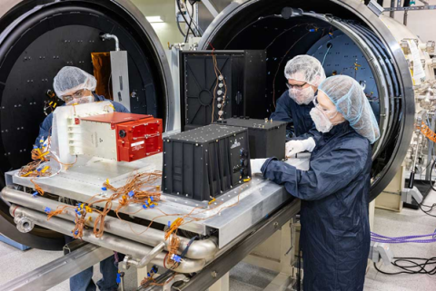 Three people wearing hairnets, masks, and protective clothing, work on a piece of electronic equipment in a laboratory setting.