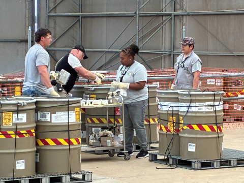 A group of employees in grey shirts move large light brown buckets with red and yellow striped tape around them inside a facility building