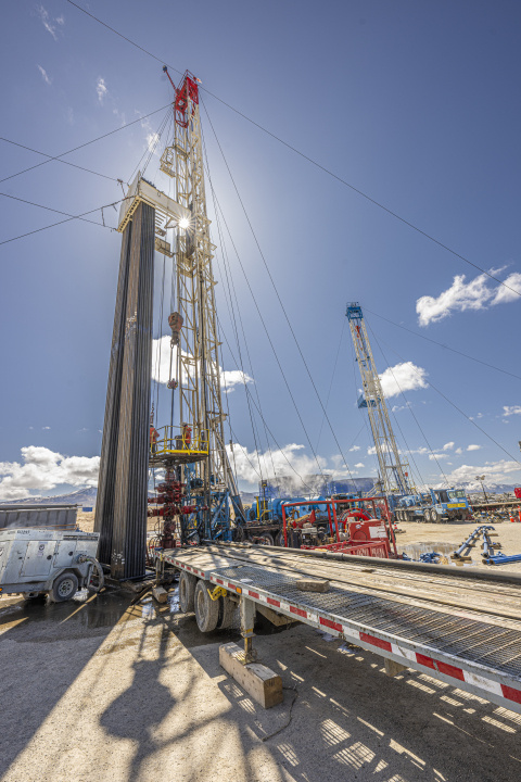 Close-up photo of a drill rig stretching up into a blue sky with the sun shining right behind the derrick (i.e. the tower).