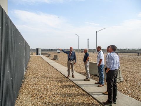 A group of professionals touring Fort St Vrain standing beside a fence on a sidewalk, one man points at the fence