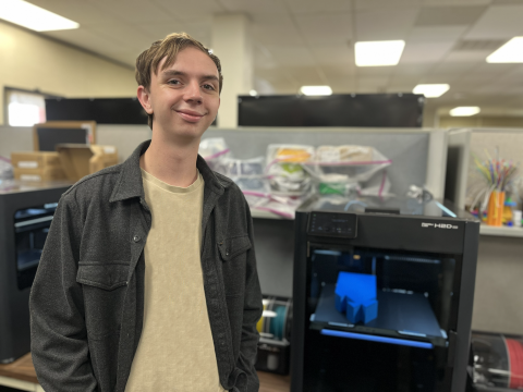 An intern in a tan shirt and black jacket smiles for the photo in front of a 3D printer