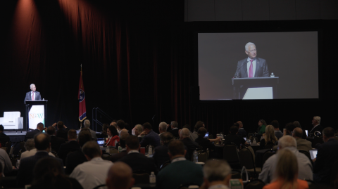 EM Assistant Secretary giving a speech, on a stage standing behind a podium, to a large crowd. To the right he is displayed on a large screen