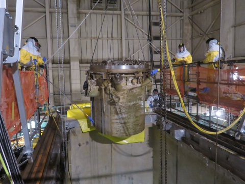 Employees in hazmat suits stand on ledges while lifting a large cylinder with ropes and wires inside a large facility building 