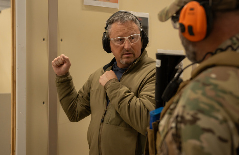 Two men at a firing range wearing camouflage speaking to each other 