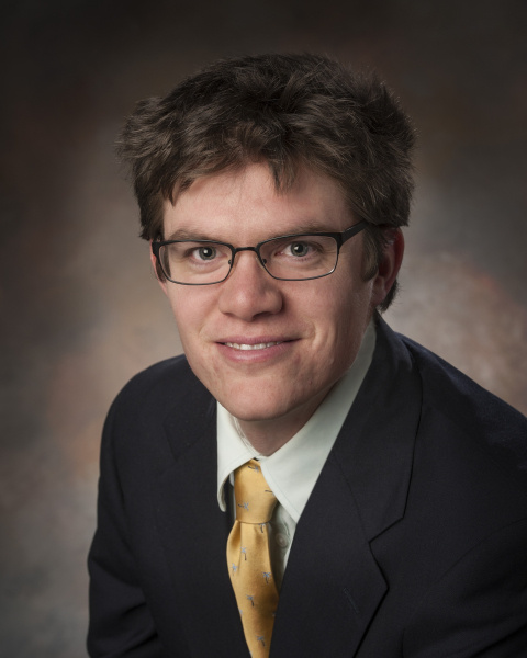 Headshot of a white man with brown hair and glasses in a black suit with gold tie