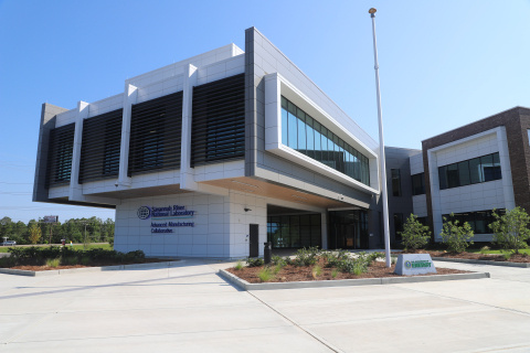 A large white building with a large overlook area in the front with lots of windows. There are plants around the building and trees in the background