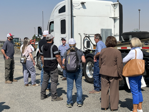 A group of people standing outside a large tractor trailer truck listening to a man talk 