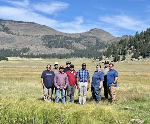 A group of individuals pose for a photo outside at the Los Alamos site with mountains and trees in the background