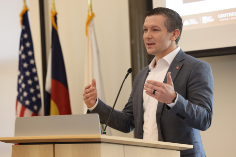 A man speaks at a lectern with American flags and the corner of a powerpoint on a large display screen in the background.