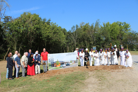 A group of people standing outside beside a dirt pile hold shovels for a groundbreaking ceremony and smile for a photo