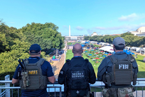 Three men facing the Washington Monument on the National Mall. On their backs are the logos/names of their agencies: NEST, the Secret Service, and the FBI.