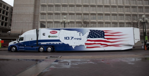 A large bus parked next to a commercial building; the bus has an image of the map of the United States and wording on the side.