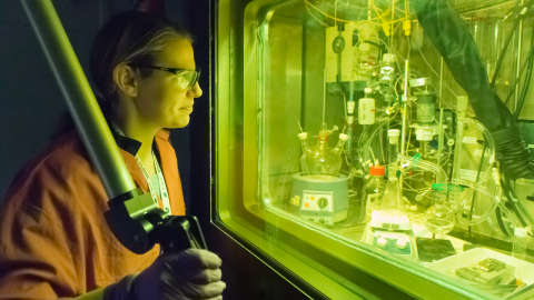A chemist operates a remote manipulator arm in a radiation-shielded cell