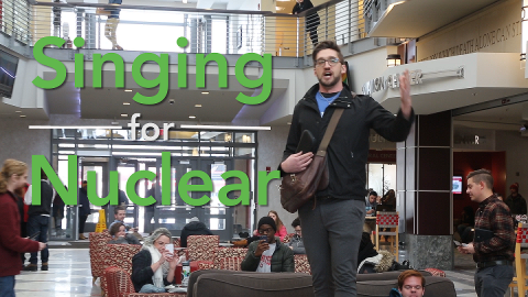 A man singing in a lobby area of a public building with the words "Opera Singer Goes Nuclear."