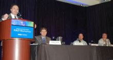 Man standing at a lectern addressing an audience, next to a long table with three people seated at it.