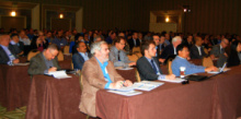 People in business attire sitting at long tables in a convention hall.
